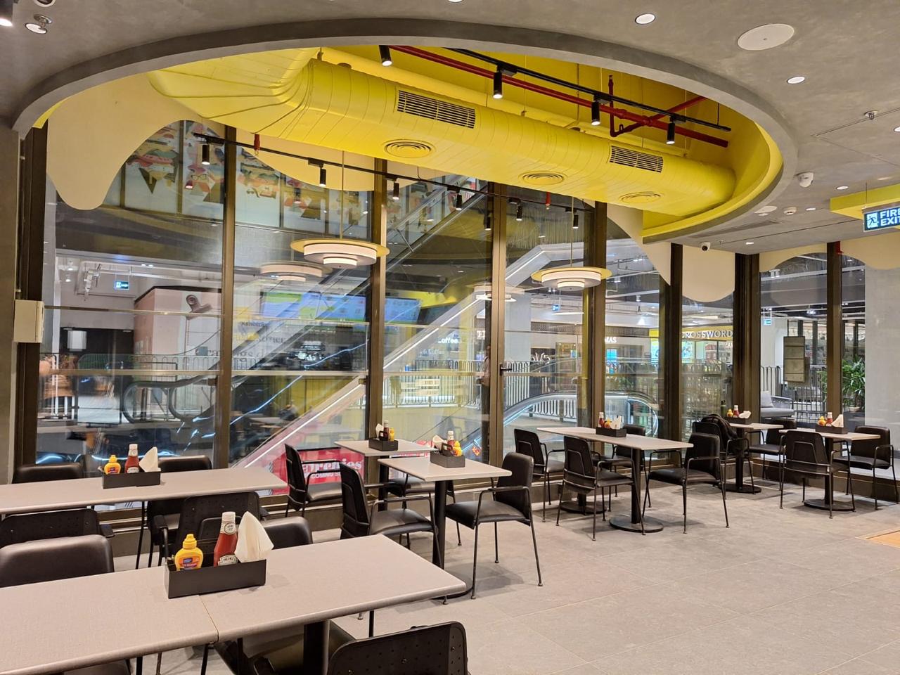 Modern indoor dining area at AKU’S DLF Midtown with empty tables, chairs, large windows, and background shops.