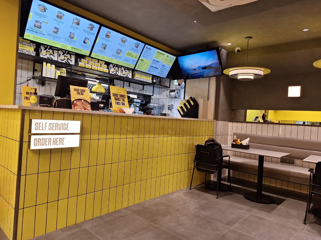 Modern fast food restaurant interior at AKU’S Punjabi Bagh with yellow tiles, digital menus, and cozy seating.
